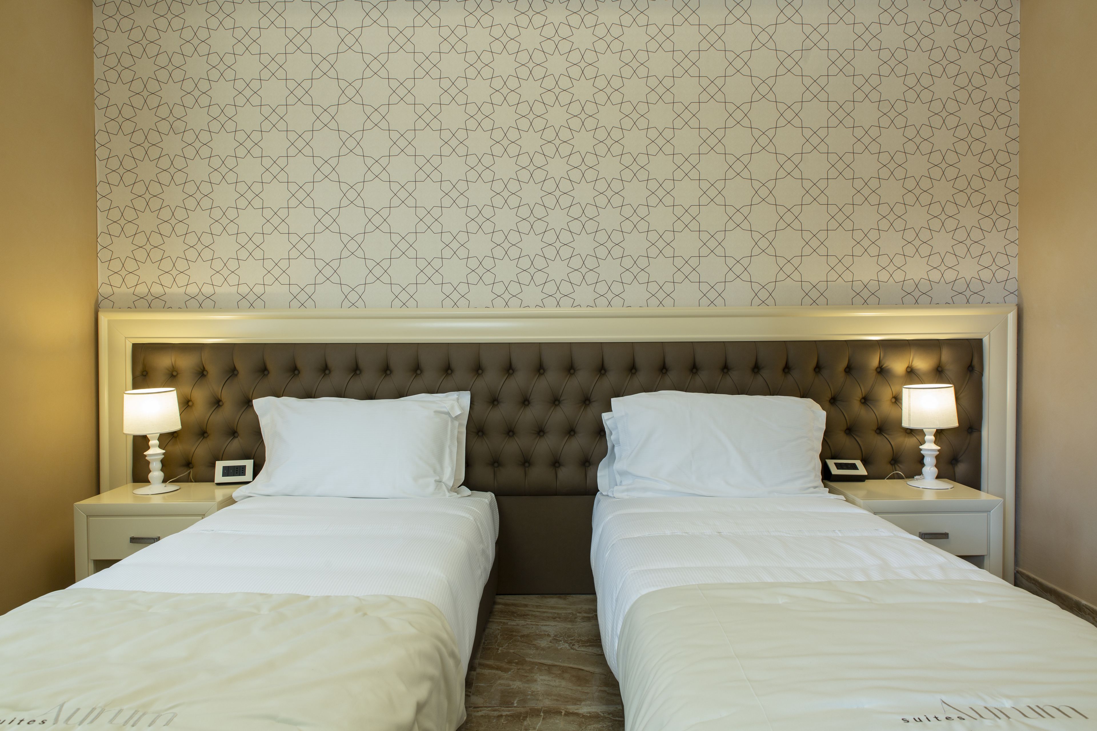 A hotel room with two beds, a tufted headboard, bedside lamps, and a patterned wallpaper.