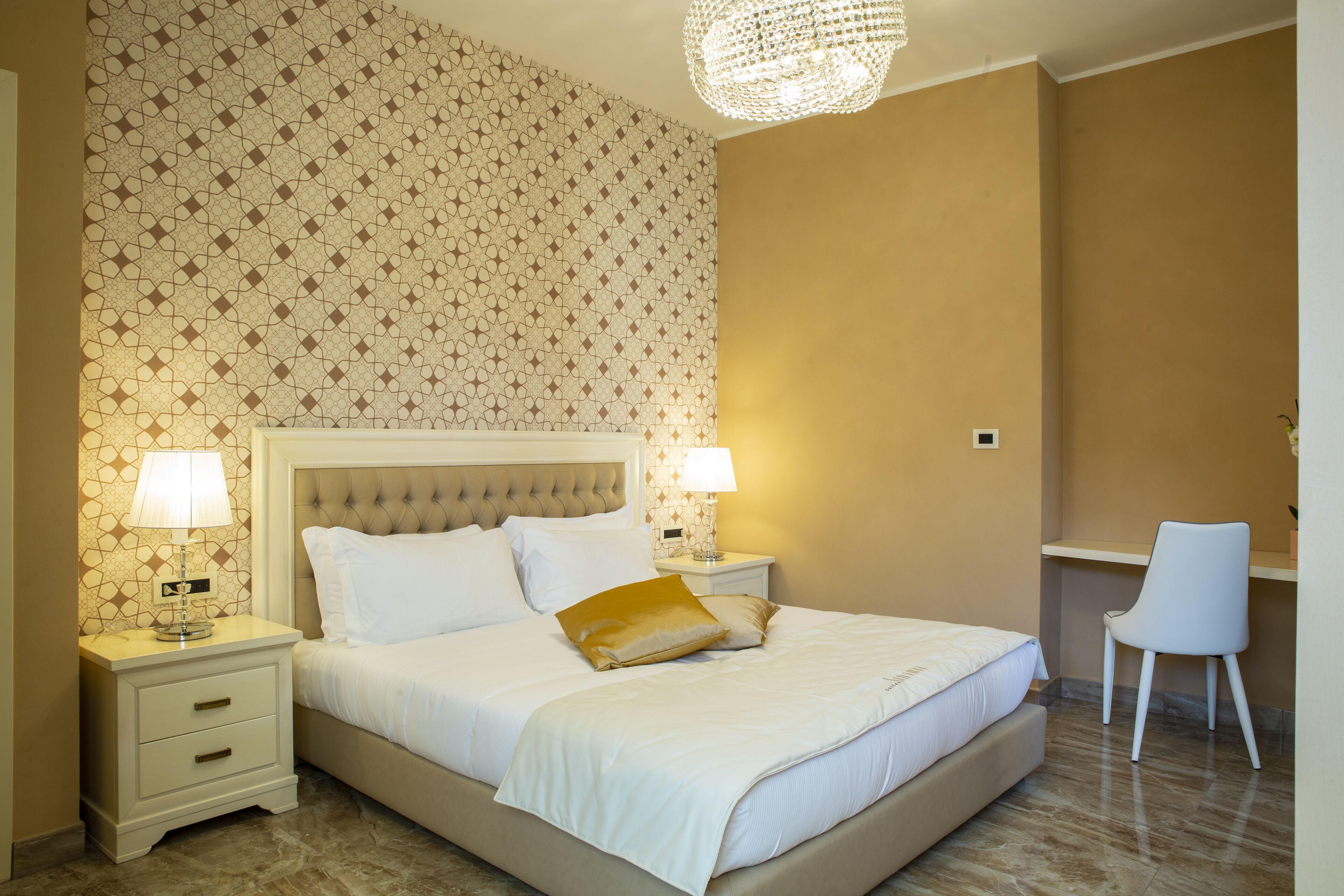 Una cama bien tendida con sábanas blancas y una única almohada dorada, flanqueada por dos mesitas de noche con lámparas, en una habitación con papel tapiz estampado y un candelabro.