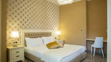 Una cama bien tendida con sábanas blancas y una única almohada dorada, flanqueada por dos mesitas de noche con lámparas, en una habitación con papel tapiz estampado y un candelabro.