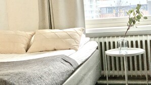 Habitación de hotel con cama, radiador, una pequeña mesa con un vaso y vistas a edificios a través de una ventana.