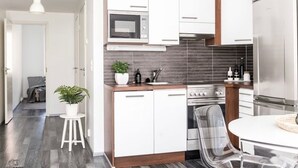 Una cocina moderna con armarios blancos, horno empotrado y microondas. Hay una zona de comedor con una mesa y sillas blancas.
