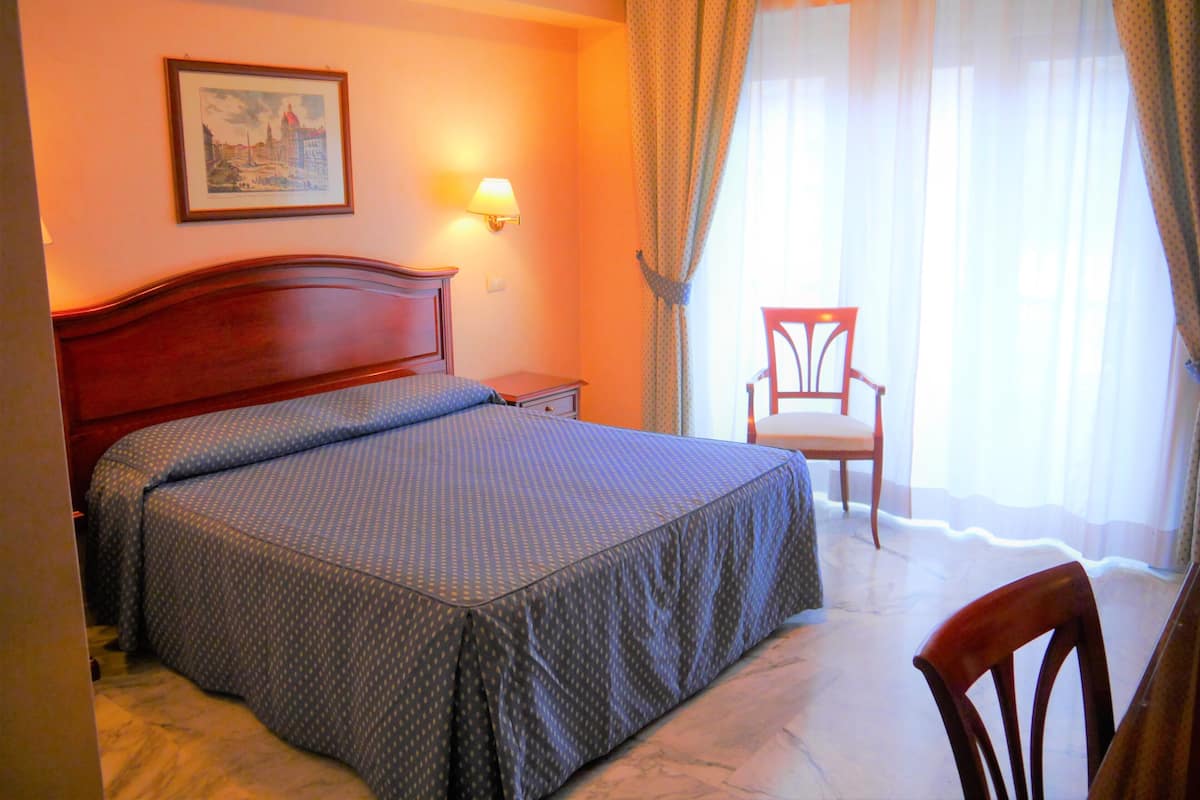 A hotel room with a large bed, a nightstand, a chair, a wall-mounted lamp, and a framed picture.