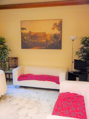 A living room with a white sofa, a red patterned throw, a painting of a building, a lamp, and two potted plants. - Hotel San Pietro (Rome)