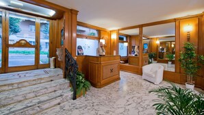 A lobby with marble flooring, wooden reception desk, white armchairs, and large windows with views of buildings outside. - Hotel San Pietro (Rome)