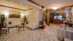 A well-lit living room with a white sofa, a coffee table, and a wooden chair. There is a painting on the wall and a chandelier on the ceiling. - Hotel San Pietro (Rome)