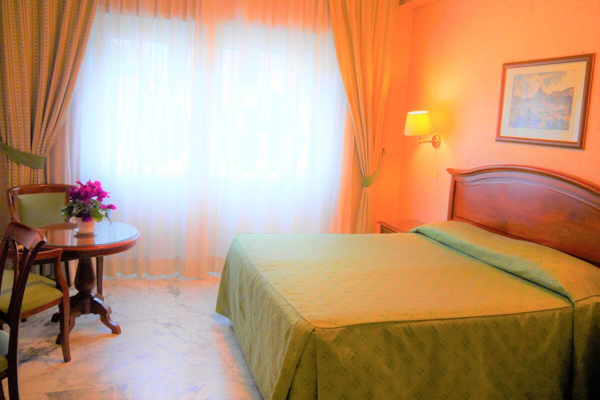 A hotel room with a bed, a chair, a small table with a vase of flowers, a lamp, and a painting on the wall.