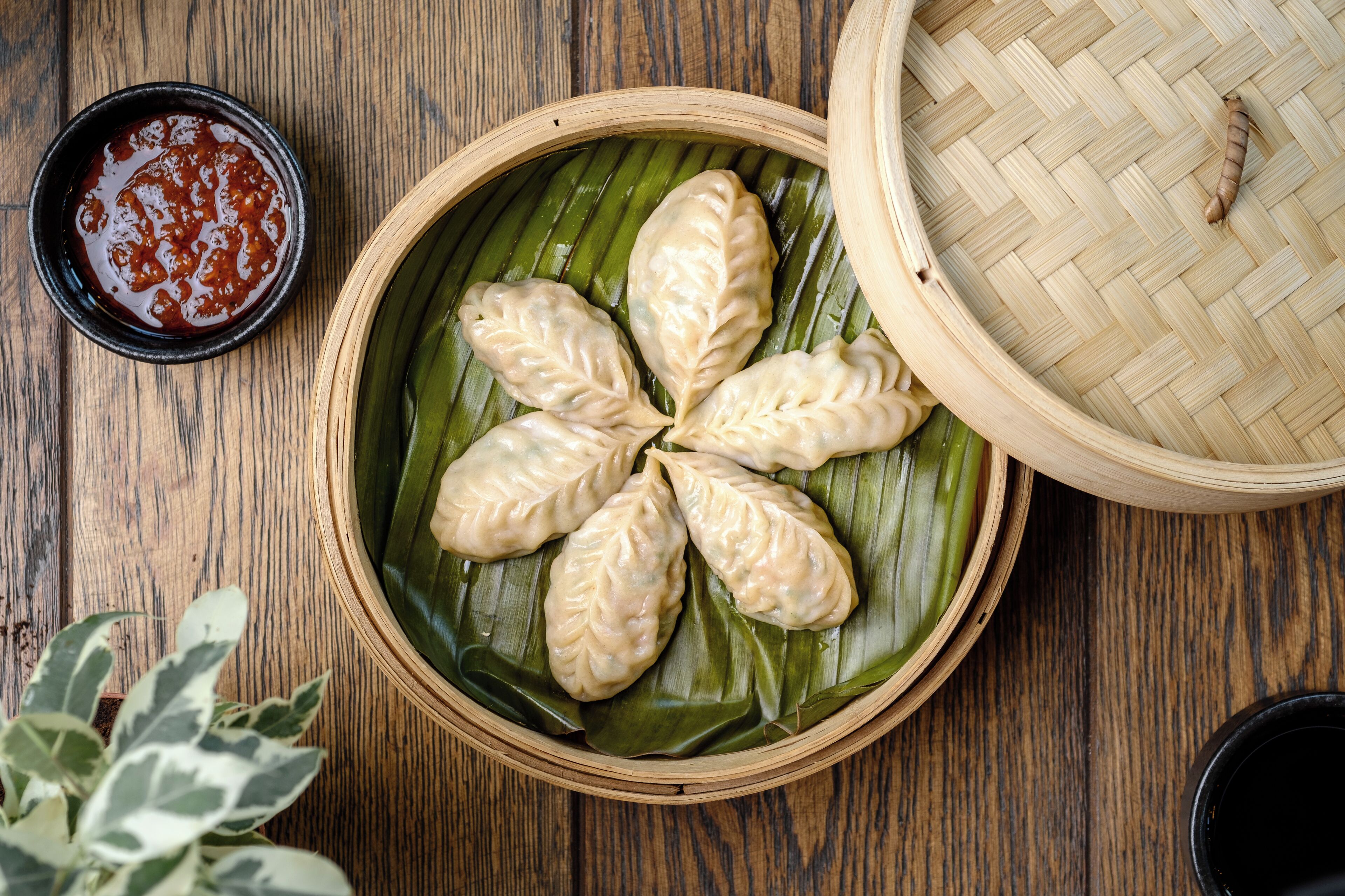 A bamboo steamer with dumplings on a banana leaf, accompanied by a small bowl of sauce.