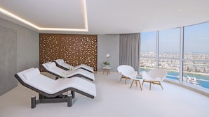Une salle de spa moderne, équipée de tables de massage, d’une petite table ornée d’un vase, et offrant une vue sur une marina grâce à de grandes fenêtres.