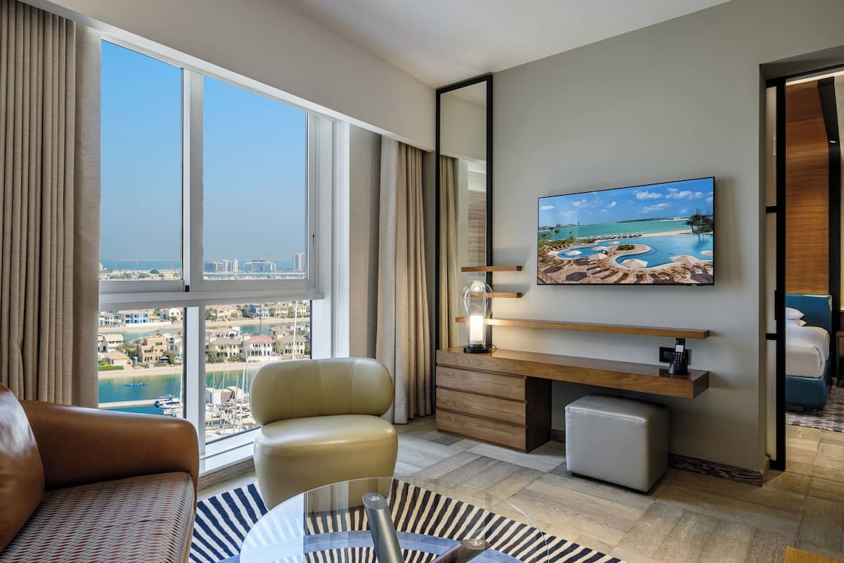 A modern hotel room with a large window offering a city view, a comfortable sofa, a desk with a computer, and a small coffee table.
