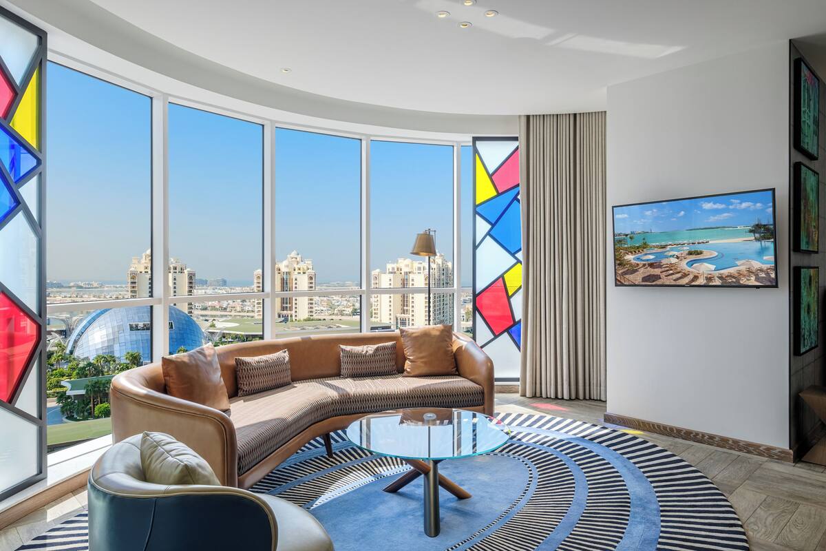 A modern living room with a curved sofa, a glass coffee table, and large windows offering a city view.