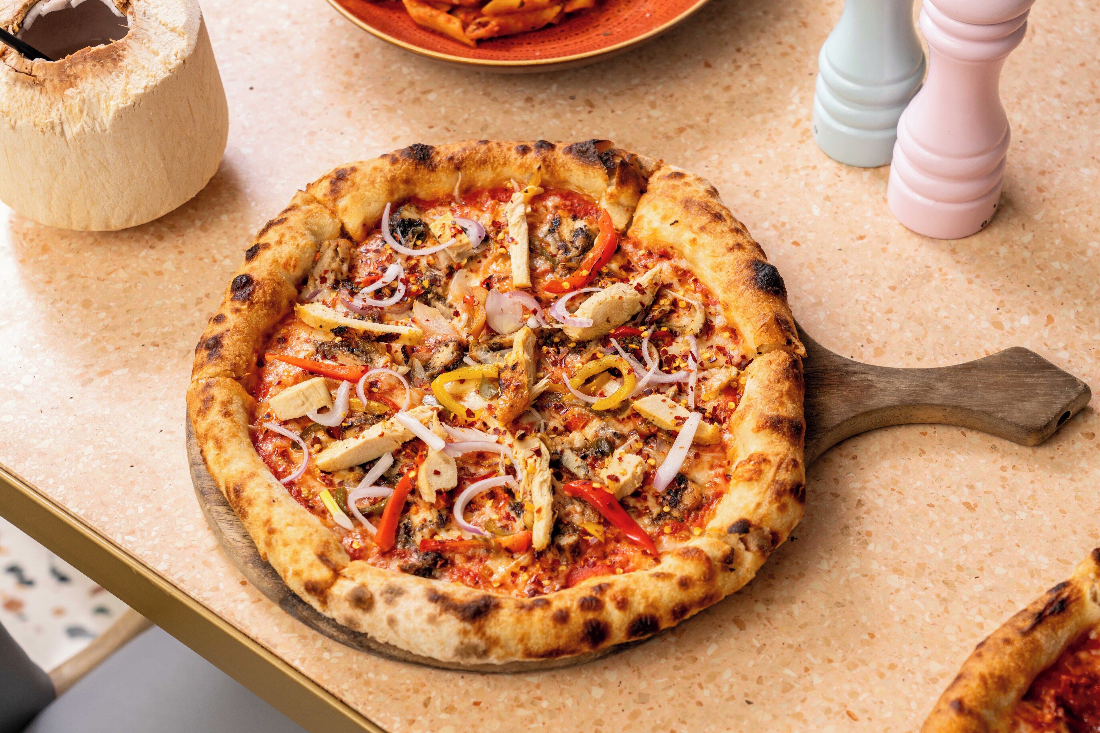 Una pizza su un tagliere di legno con un manico, circondata da un piano di lavoro maculato.