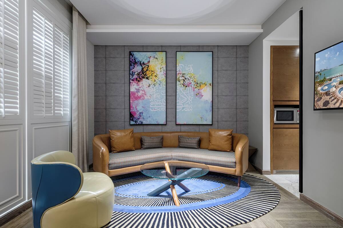 A modern living room with a curved sofa, a glass coffee table, and a striped rug. There are two abstract paintings on the wall and a television on the right.