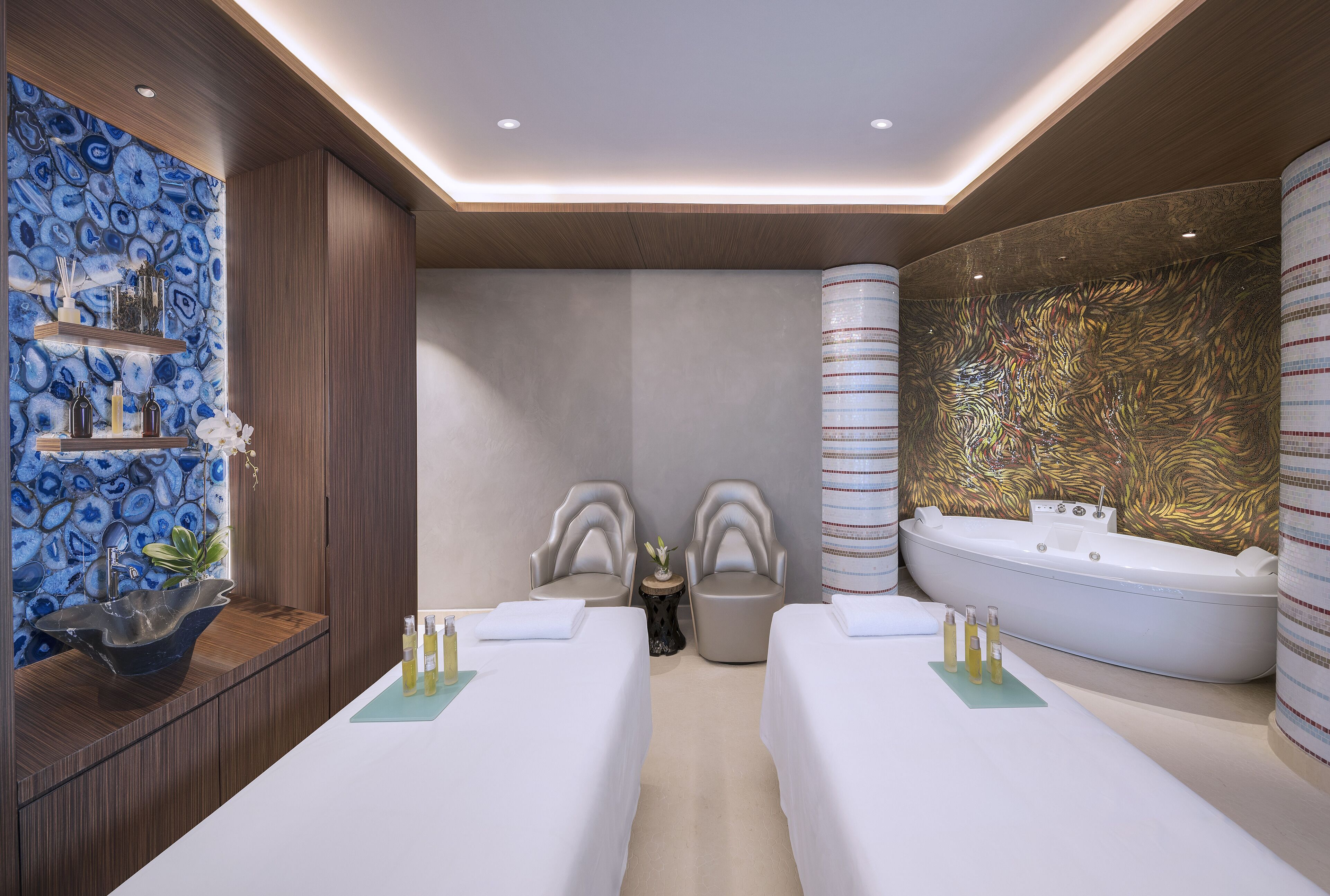 A modern spa room with two massage tables, a large bathtub, and a decorative wall with a blue and gold pattern.