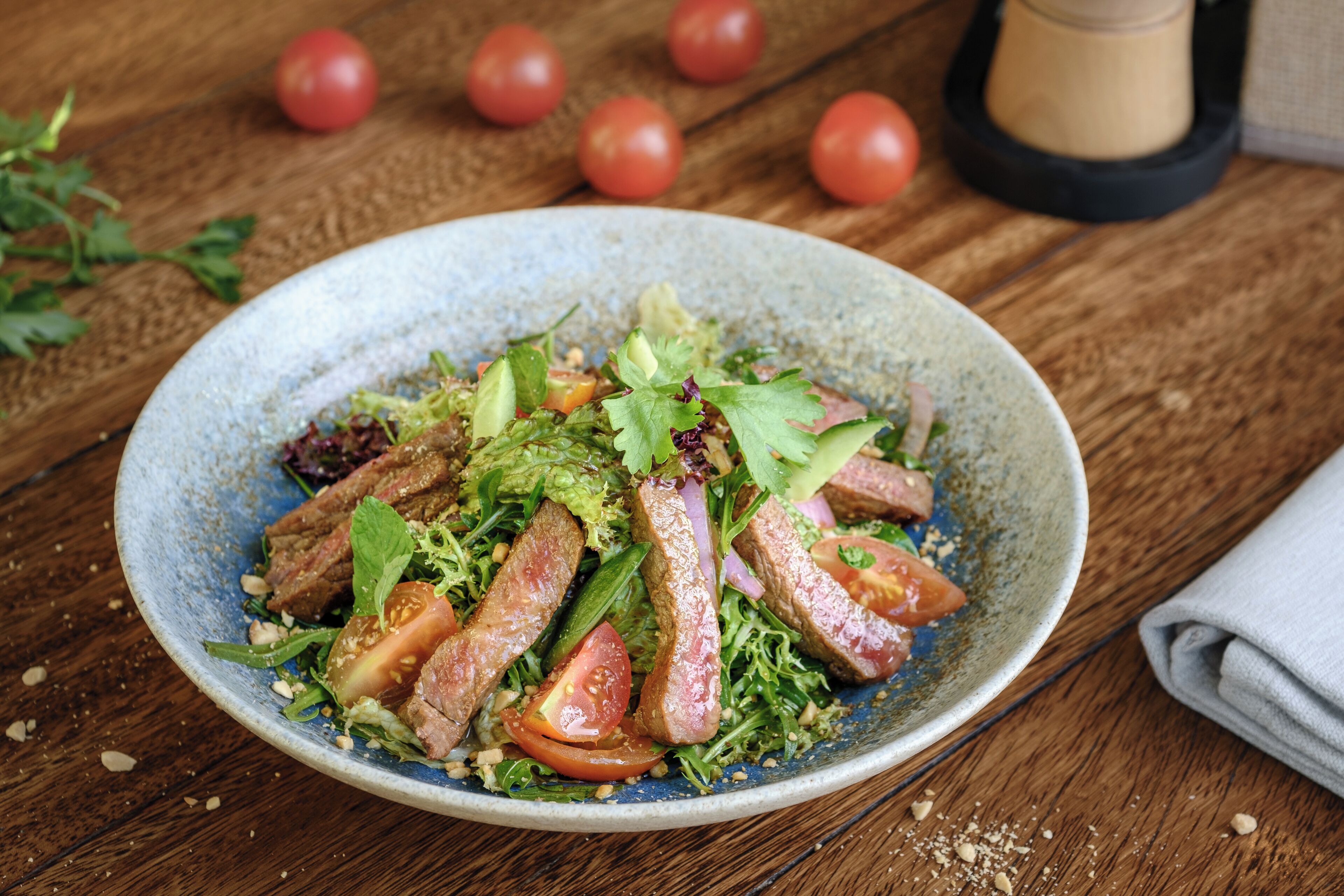A plate of food with meat, vegetables, and a blue bowl on a wooden table.