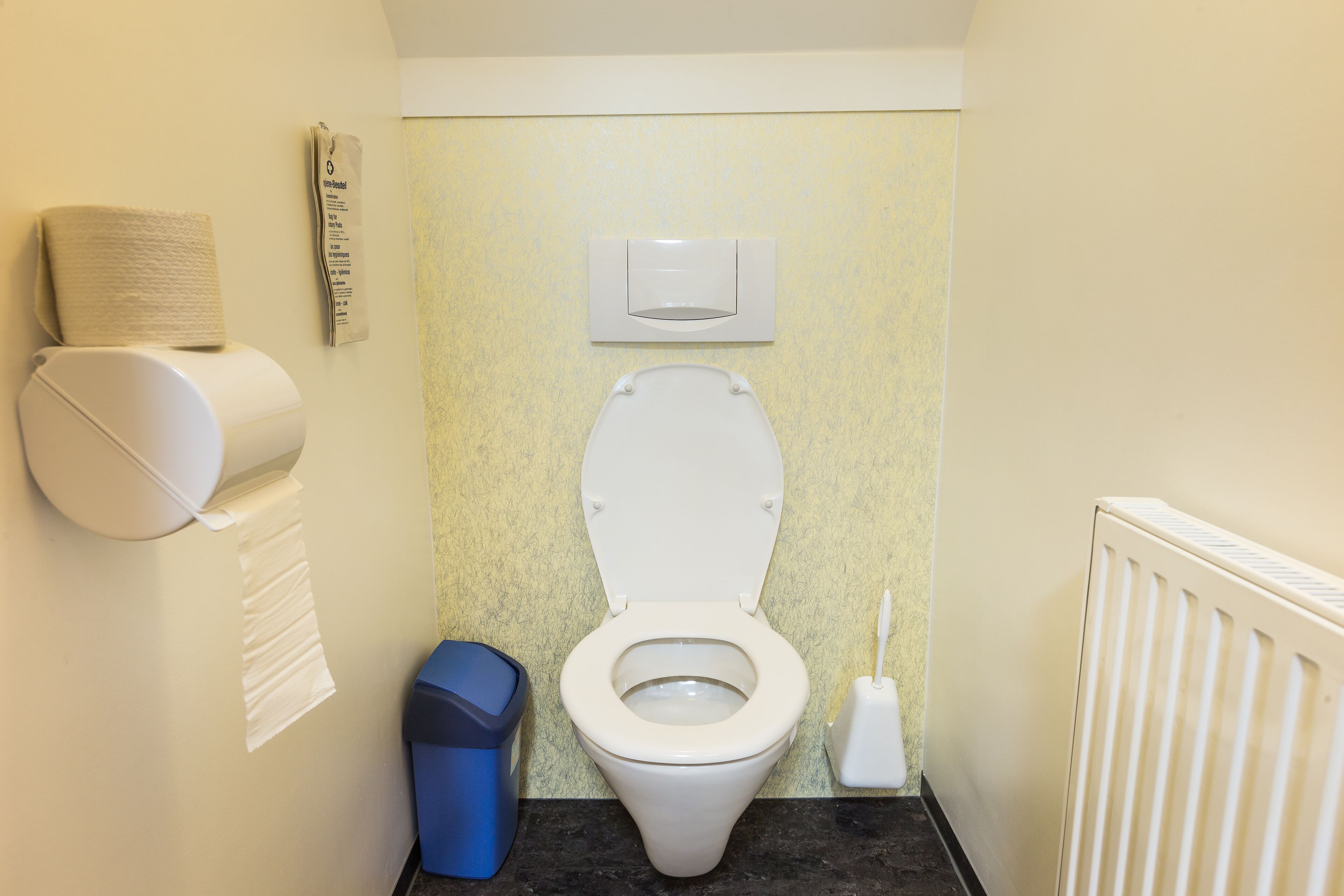 A small bathroom with a toilet, a trash bin, a paper towel dispenser, and a radiator.