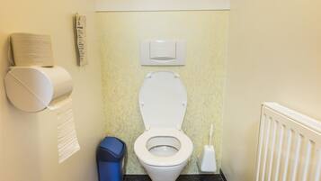 A small bathroom with a toilet, a trash bin, a paper towel dispenser, and a radiator.