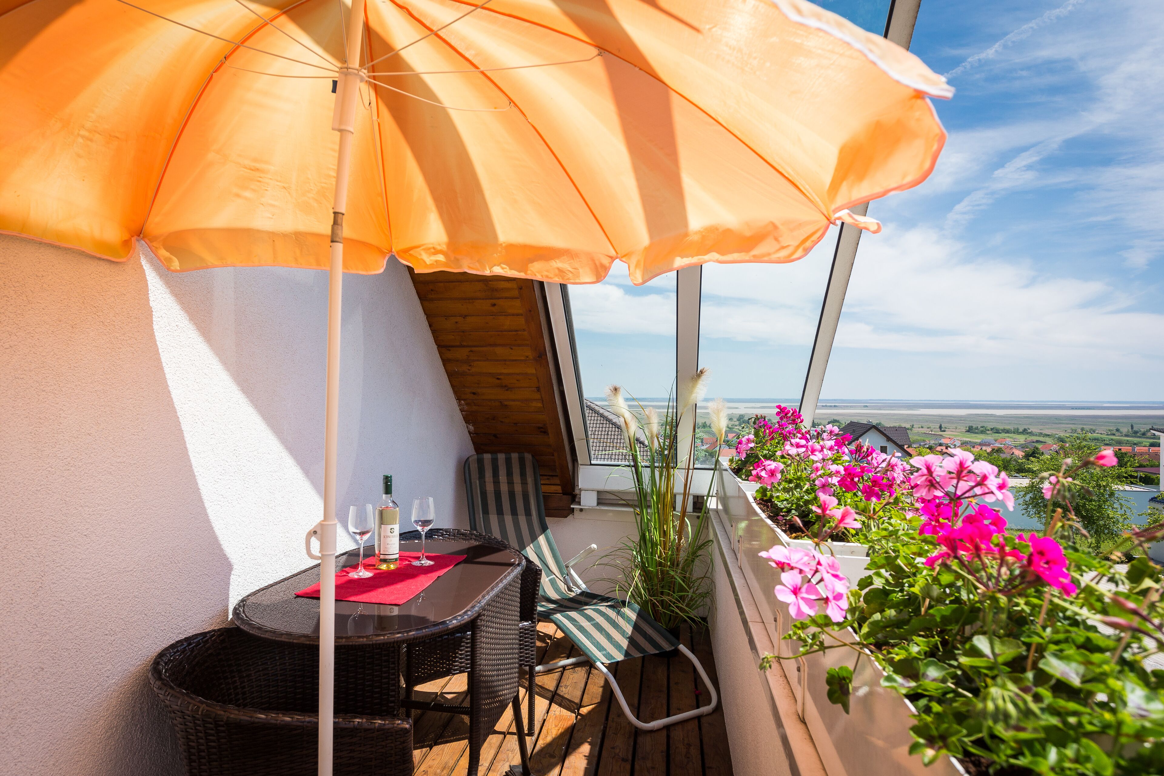 Varanda com mesa e cadeiras de vime, um guarda-sol laranja e flores cor-de-rosa.