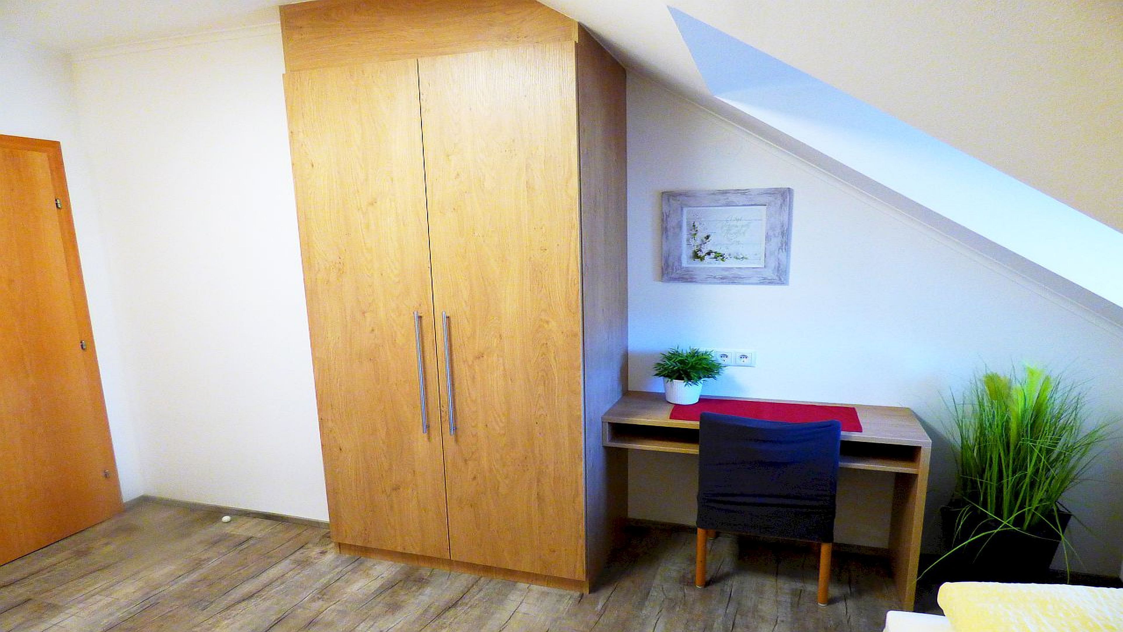 Quarto com guarda-roupa de madeira, escrivaninha com cadeira, uma pequena planta e um quadro na parede.