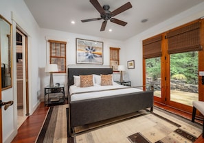 A bedroom with a bed, a ceiling fan, a nightstand, a lamp, a window with blinds, and a view of the outdoors.