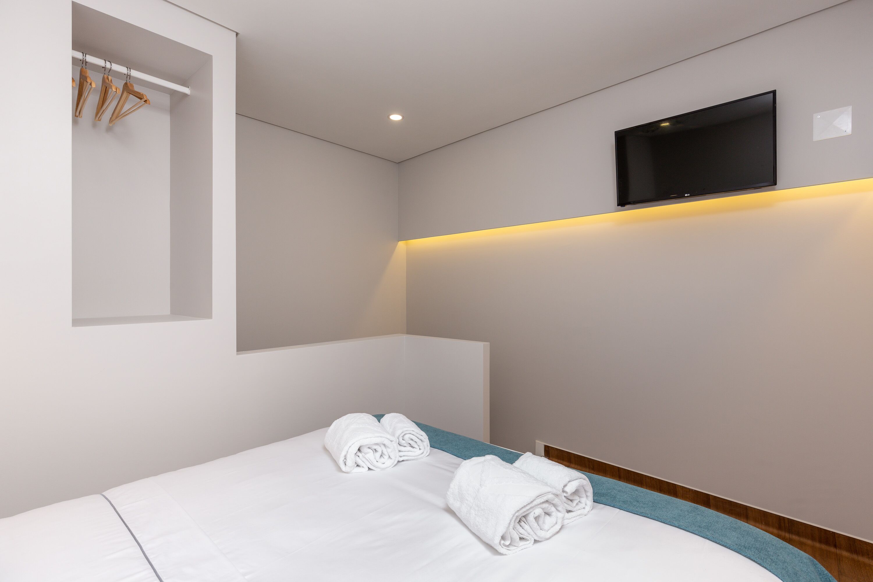 A hotel room with a bed, a flat-screen TV mounted on the wall, and a built-in shelf with towels.