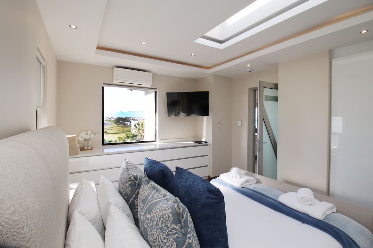 A modern hotel room with a bed, a sofa, a TV mounted on the wall, and a window with a view of the outdoors.