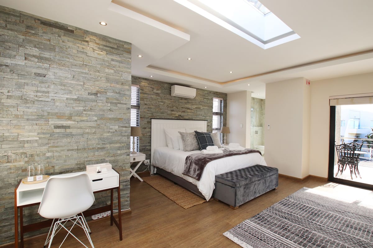 A modern bedroom with a skylight, a bed with a grey upholstered bench, a desk with a chair, and a view through a glass door.