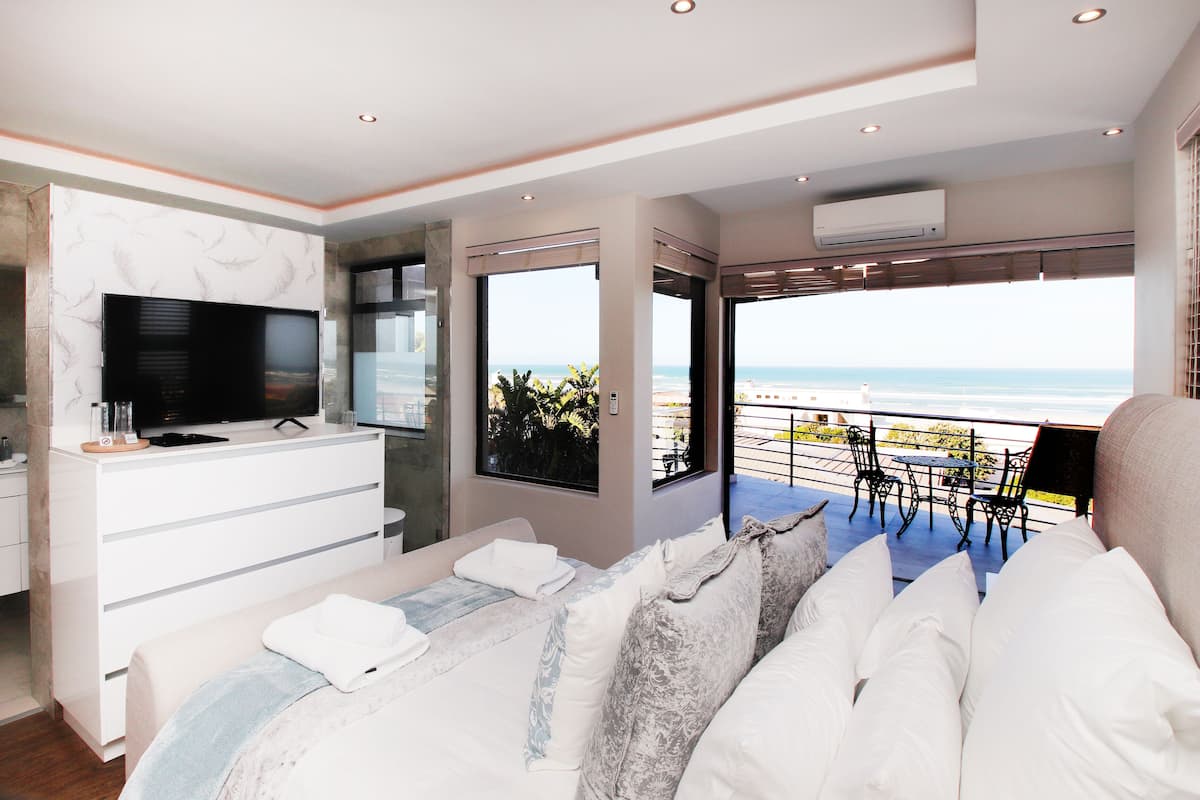 A modern bedroom with a large bed, a TV, and a balcony with a view of the beach.