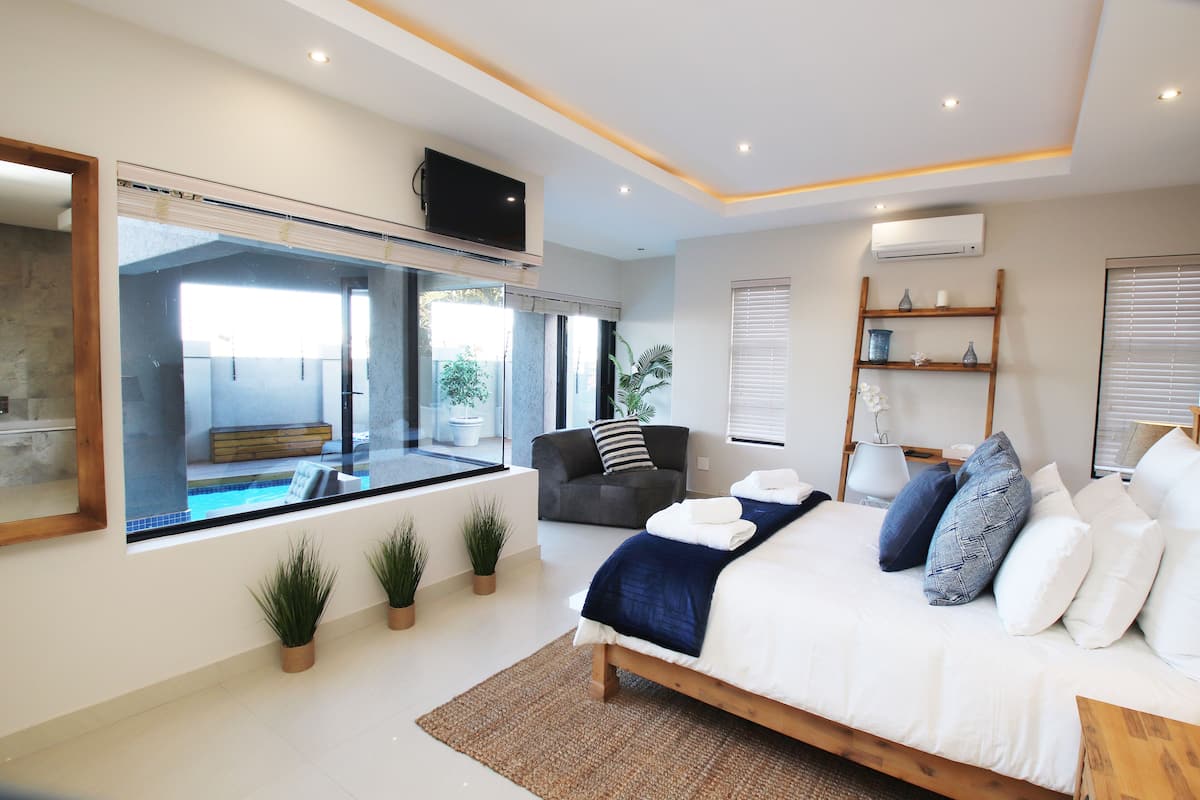A modern bedroom with a large bed, a TV mounted on the wall, a window with a view of a pool, and a small shelf with decorative items.