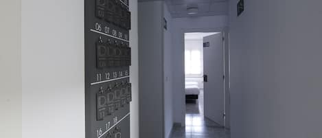 Pasillo de hotel con puertas numeradas que dan a habitaciones, una cama visible a lo lejos y piso de baldosas.