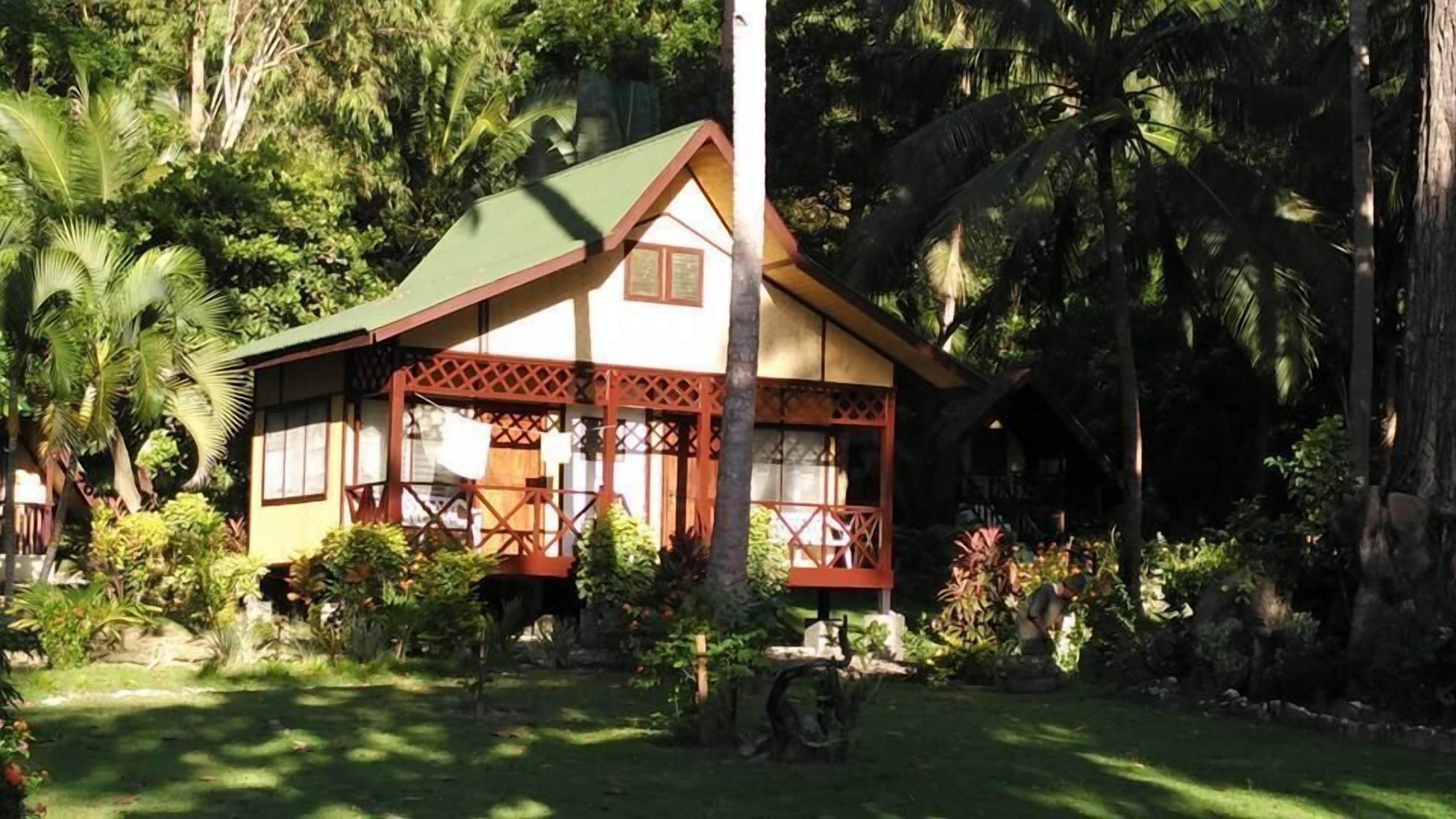 A small, single-story house with a green roof and a porch, surrounded by lush greenery.