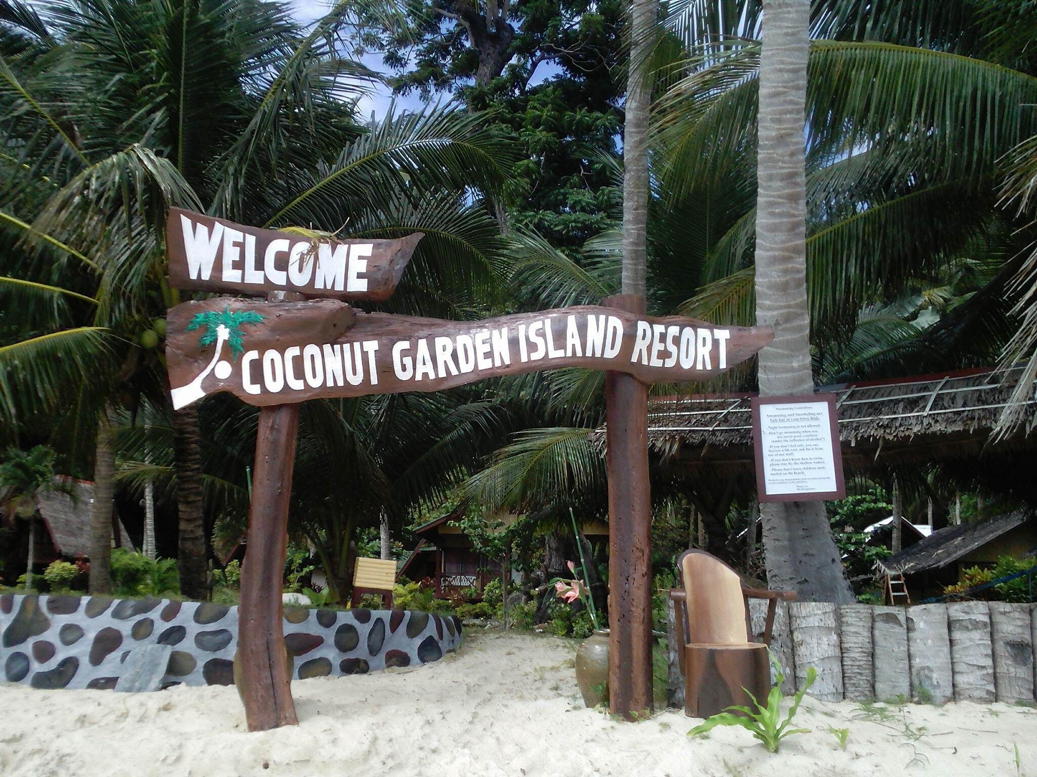 A wooden sign with 'WELCOME' and 'Coconut Garden Island Resort' written on it, surrounded by palm trees and a sandy area.