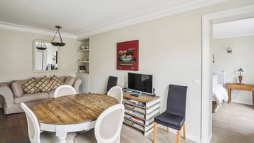 A living room with a round wooden table, white chairs, a sofa, a television, and a bookshelf.