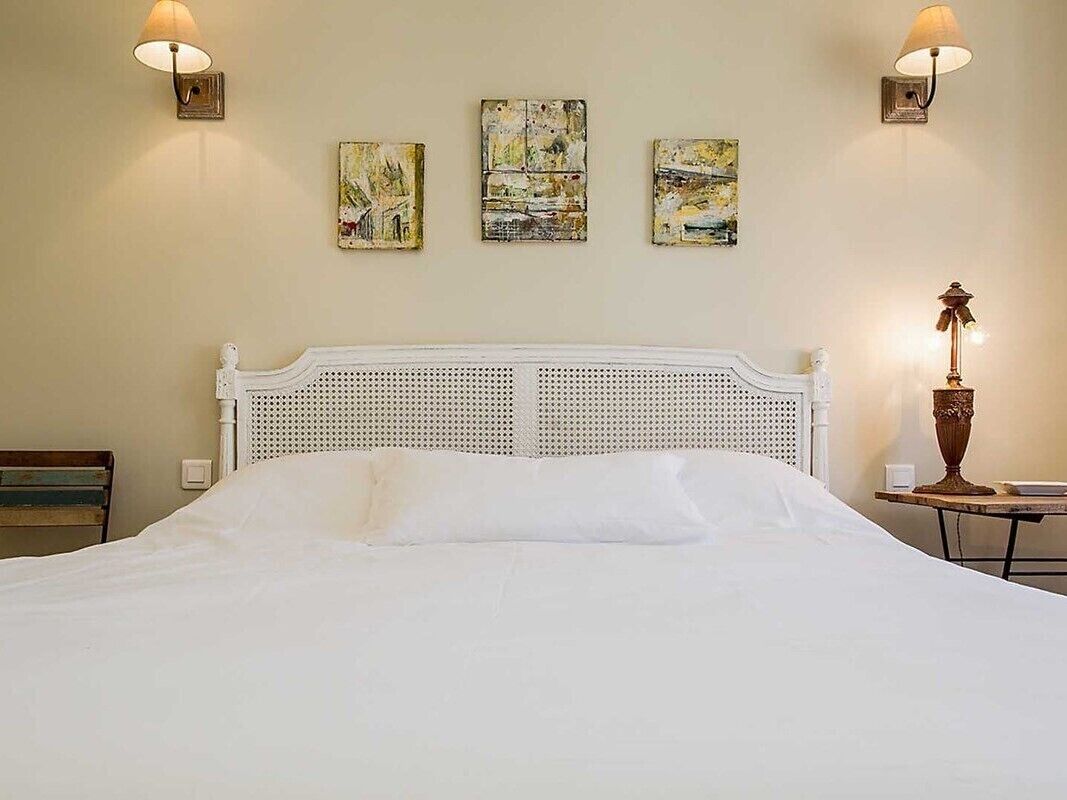 Un lit bien fait, avec une tête de lit blanche, deux lampes fixées au mur et trois tableaux abstraits accrochés au mur.