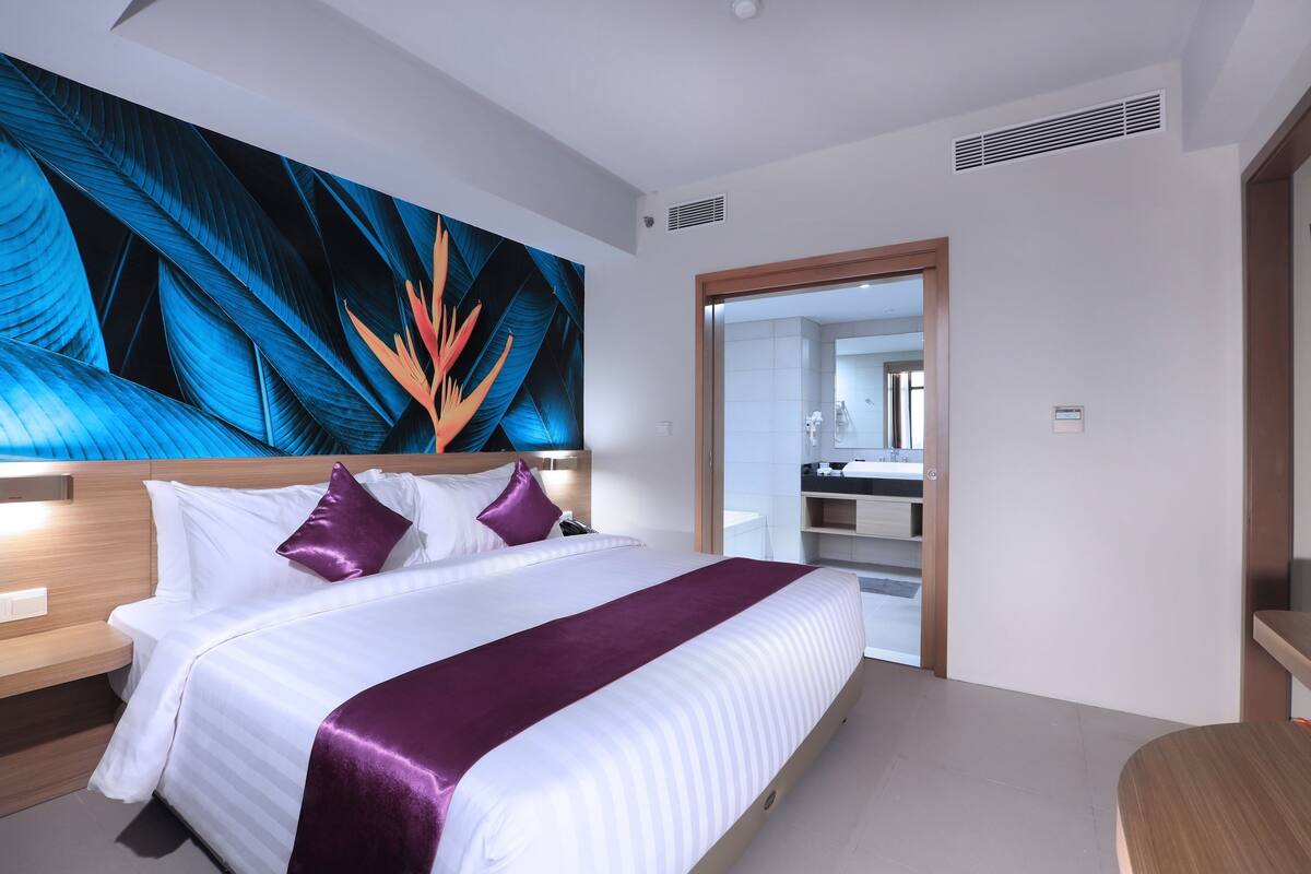 A modern hotel room with a large bed, a headboard with built-in lighting, a large wall mural, and a view into the bathroom.