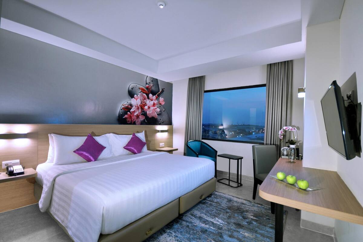 A modern hotel room with a large bed, a desk with a chair, a TV, and a view of the city at night.