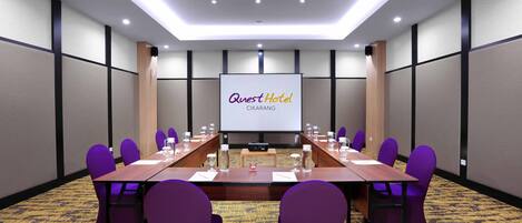 A conference room with a long table, purple chairs, and a screen displaying "Quest Hotel Cikarang".