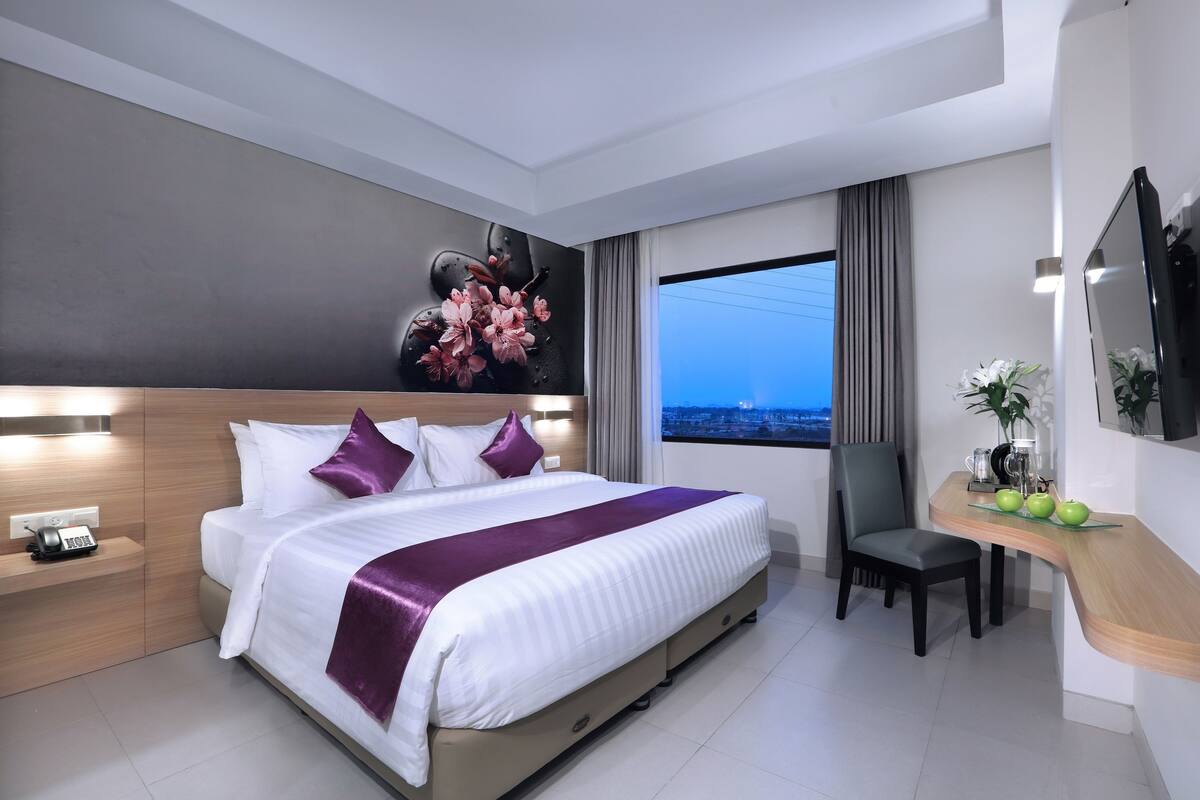 A modern hotel room with a large bed, a desk with a chair, a TV, and a window with a city view.