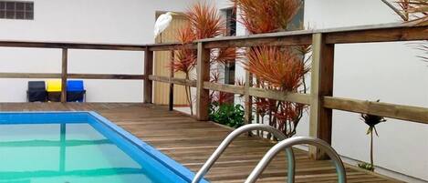 A pool area with a wooden deck, a metal pool ladder, and a white wall with a window and air conditioning unit.