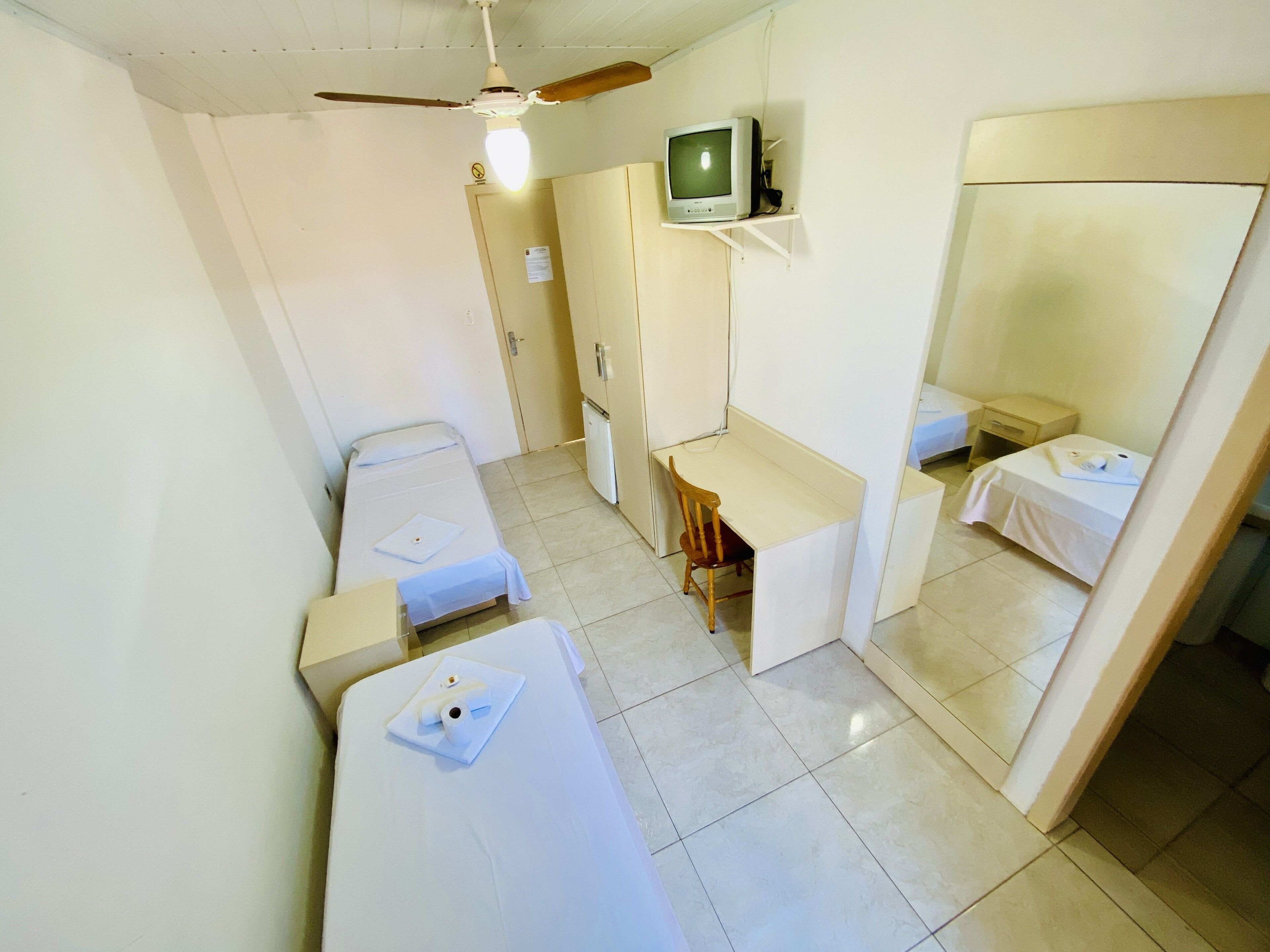Habitación de hotel compacta con dos camas individuales, un escritorio con silla, un televisor pequeño y un ventilador de techo.
