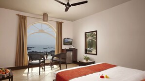 A hotel room with a bed, a desk, a chair, a small table with fruit, a potted plant, and a view of the ocean. - O Pescador (Panaji)