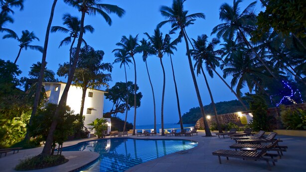 A pool area with lounge chairs, palm trees, and a building with a white facade. - O Pescador (Panaji)