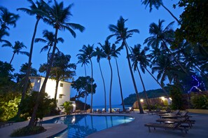 A pool area with lounge chairs, palm trees, and a building with a white facade. - O Pescador (Panaji)