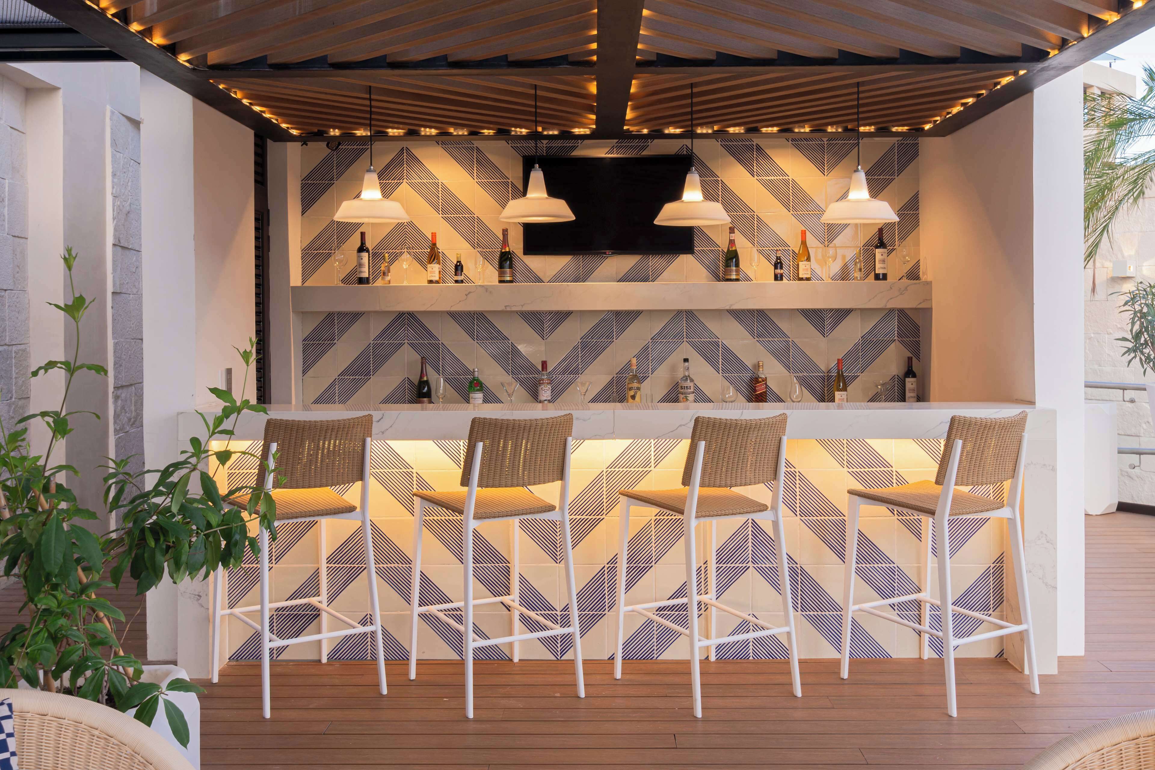 A bar area with a geometric patterned wall, white bar stools, and pendant lighting.