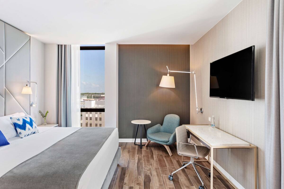 Habitación de hotel moderna con cama, escritorio, silla, televisor y vista a la ciudad.