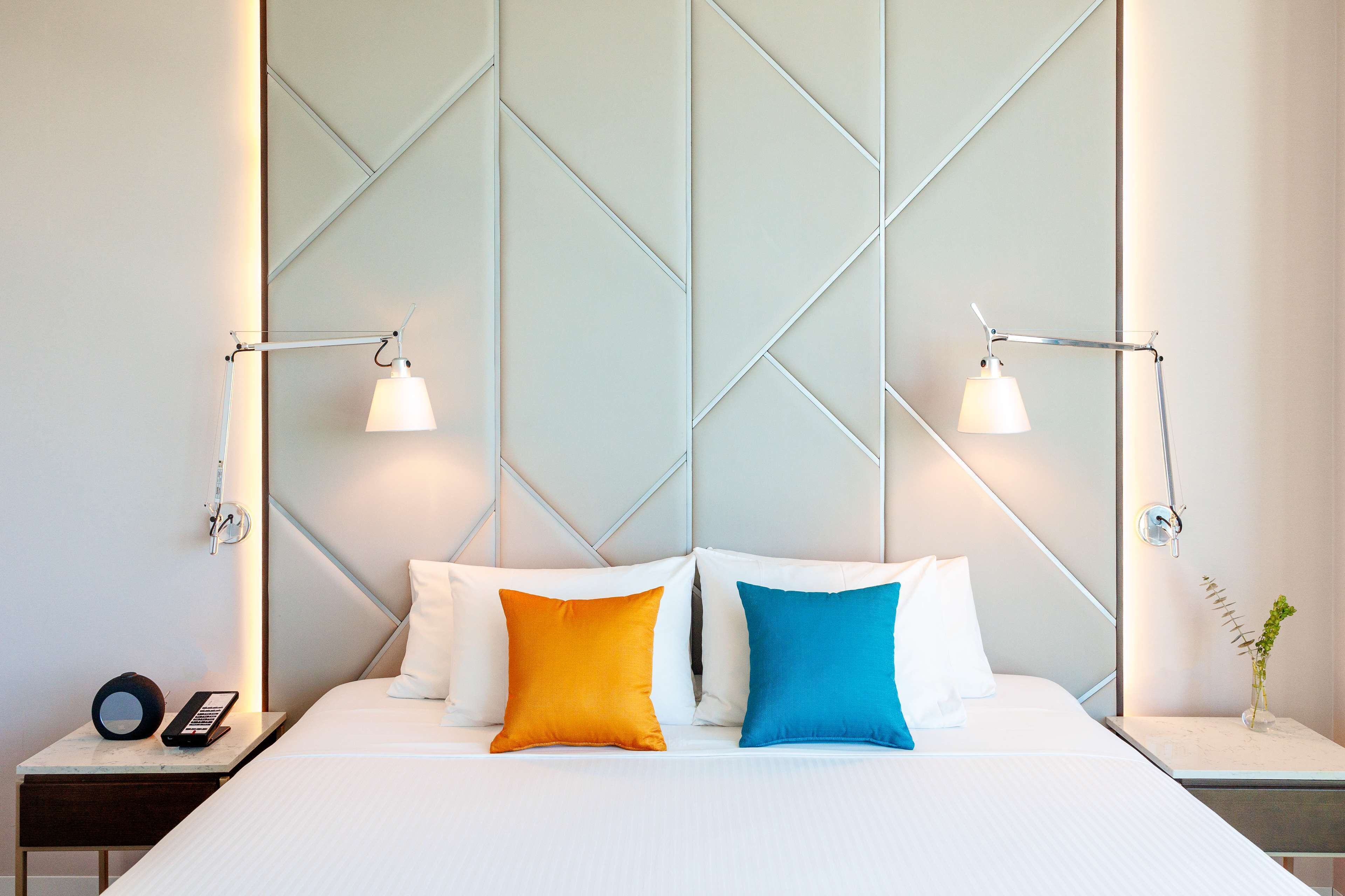 A neatly made bed with white and two brightly colored pillows, a bedside table with a lamp, and a small vase with a plant.