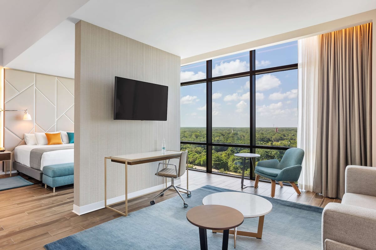 Una habitación de hotel moderna con un ventanal grande, un televisor de pantalla plana, un escritorio con una silla, un sofá y una mesita.
