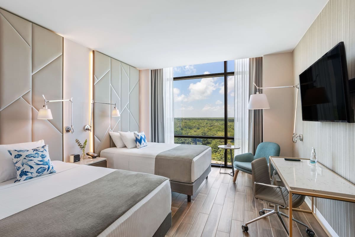 Habitación de hotel con dos camas, un escritorio, una silla, un televisor y un amplio ventanal con vistas al paisaje.
