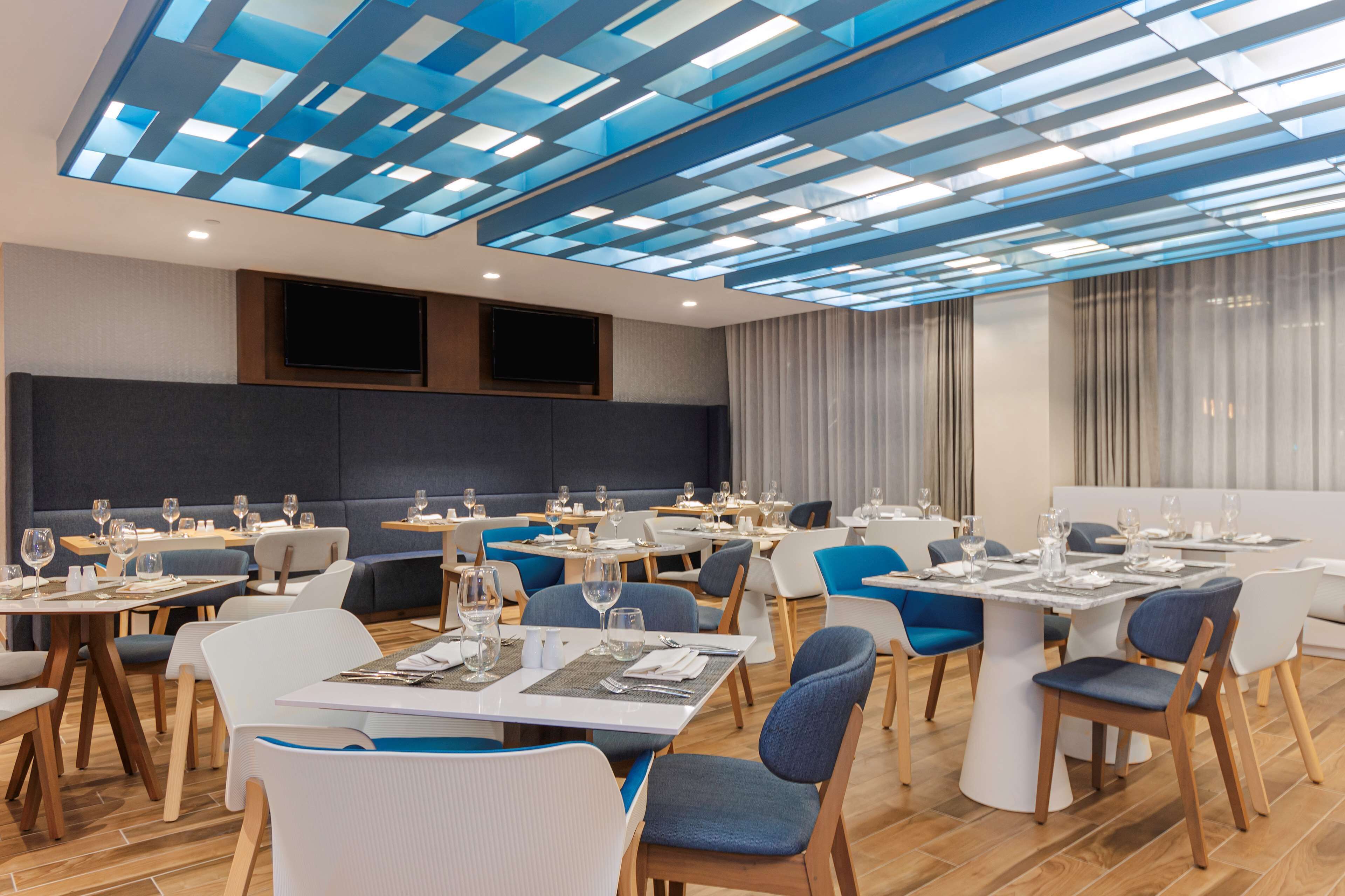 A modern dining area with multiple tables and chairs, a flat-screen TV mounted on the wall, and a ceiling with a geometric pattern.