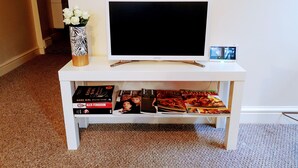 A white entertainment center with a flat-screen TV, a small digital photo frame, and a vase with white flowers. Below the TV, there are books and magazines on a shelf. - The Hazeldene Apartment (Cardiff)
