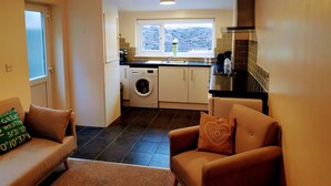 A compact living space with a sofa, a coffee table, a washing machine, a kitchen with a window, and a ceiling light. - The Hazeldene Apartment (Cardiff)